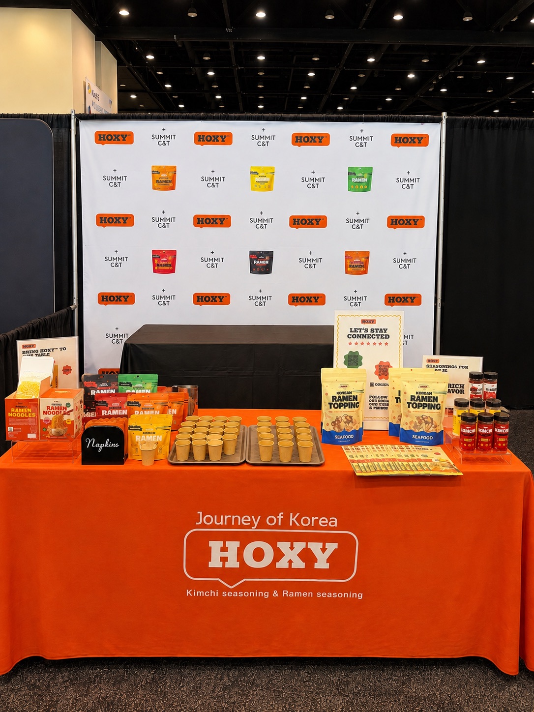 HOXY booth at the KeHE show: step-and-repeat backdrop, orange table cover and sampling setup